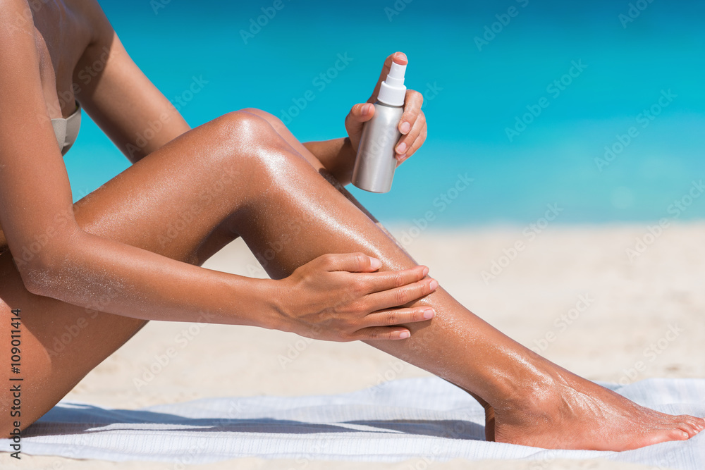 Sunscreen suntan lotion in spray bottle. Young woman in spraying tanning  oil on her leg from bottle. Lady is massaging sunscreen lotion while  sunbathing at beach. Female model during summer vacation. Photos
