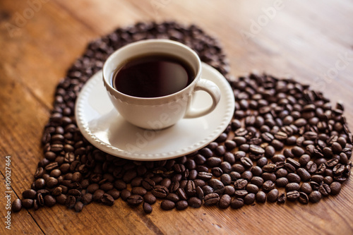 Heart from coffee beans with cup of coffee on wooden background