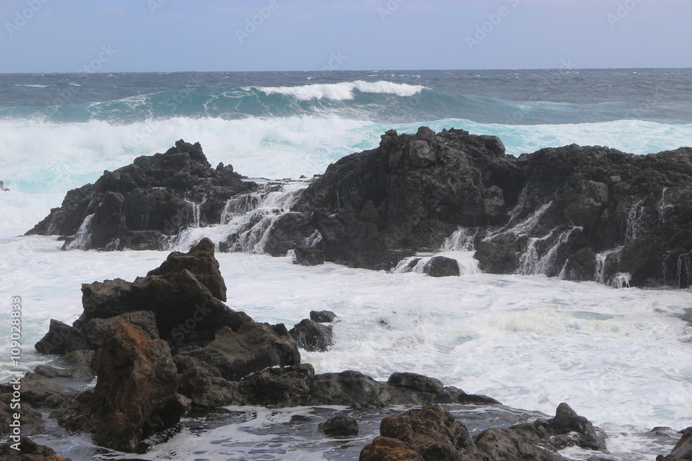 Wild sea with breaking waves on black lava rocks on the east coast of Lanzarote, Canary Islands, Spain. In the Northeast of the Island between Arrieta and Orzola.. Wild sea with breaking waves on black lava rocks on the east coast of Lanzarote, Canary Islands, Spain. In the Northeast of the Island between Arrieta and Orzola..