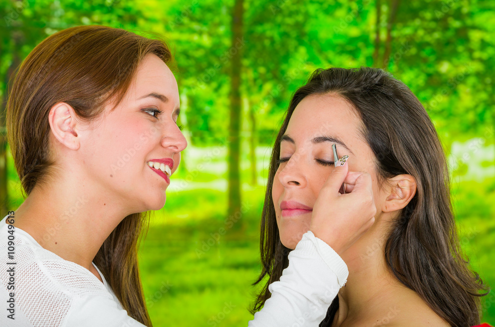 Woman performing eyebrow hair removal using tweezers on clients face, uncomfortable facial expressions, green garden background Woman performing eyebrow hair removal using tweezers on clients face, uncomfortable facial expressions, green garden background
