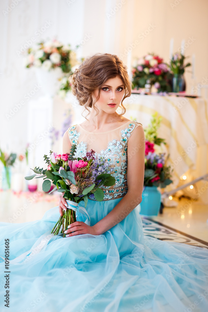 Beautiful young lady in a luxury blue dress in elegant interior with a bouquet of flowers