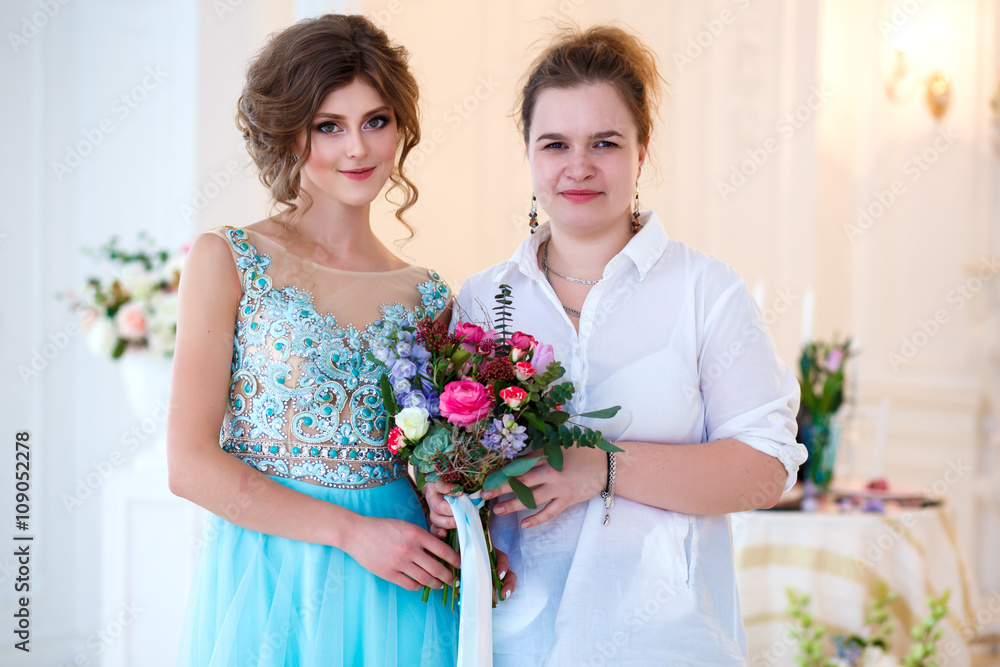 Beautiful young lady in a luxury blue dress in elegant interior with a bouquet of flowers with a florist woman