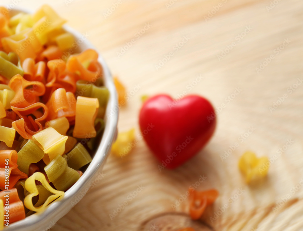 Dry multicolored heart shaped pasta in white bowl on wooden table