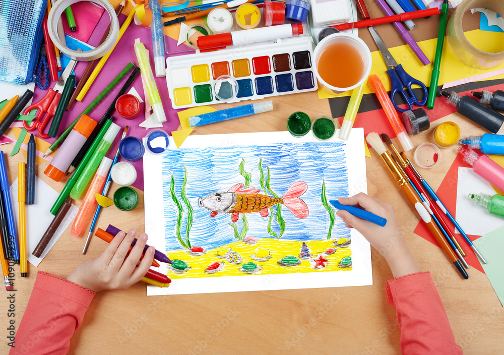 Fototapeta premium child drawing fish underwater and seabed, top view hands with pencil painting picture on paper, artwork workplace