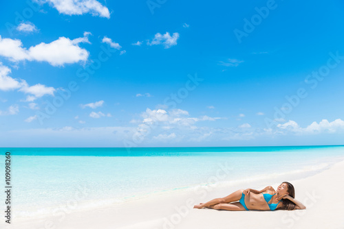 Sexy bikini suntan tan woman relaxing sun tanning on beach vacation luxury resort paradise getaway lying down on perfect white sand sunbathing in tropical Caribbean travel destination. Sky copyspace.