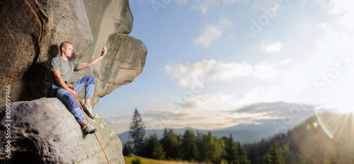 Climber is sitting on the big natural boulder secured with the rope against blue sky and mountains. Young man is taking picture by the smart phone at the mountains. Summer. Copy-space on the right