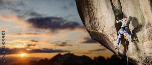 Young athletic male rock climber falling down from challenging route on overhanging cliff with rope and carbines on rocky wall against scenic sunset background. Summer time. Panoramic image