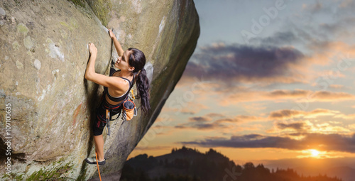 Fototapeta Young attractive female rock climber climbing challenging route on steep rock wall against scenic sunset background