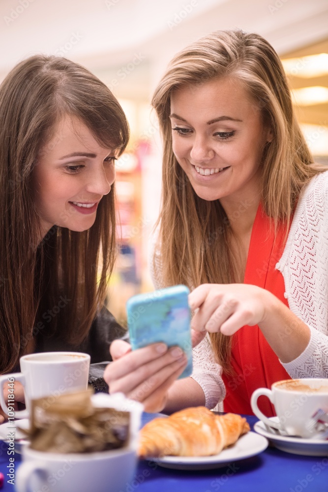 Two women looking at mobile phone while having snacks and coffee in coffee shop Two women looking at mobile phone while having snacks and coffee in coffee shop