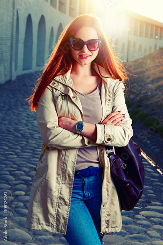 Street fashion portrait of young foxy-haired woman in a jeans and violet sunglasses with a leather handbag against the setting sun. Cold season. Pink lips. Warm sunset colors.