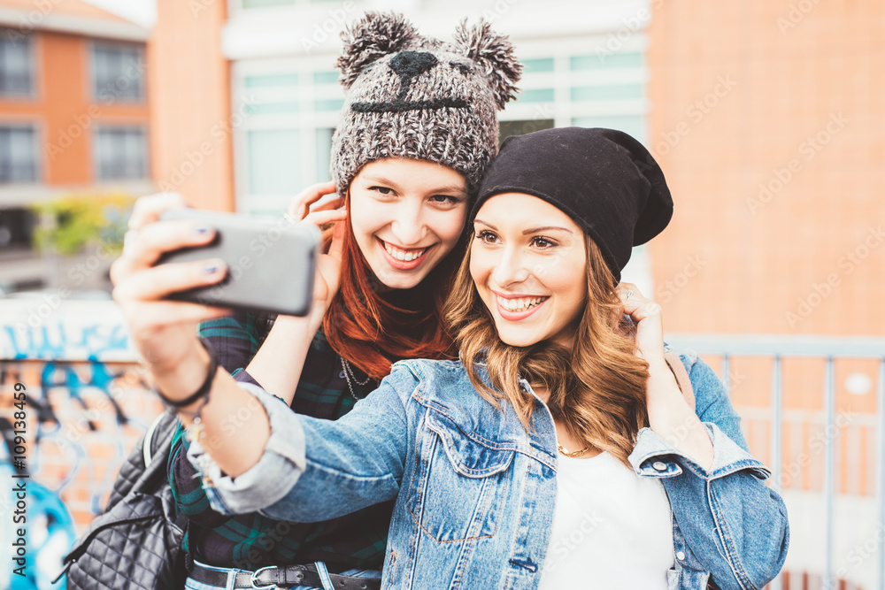 Half length of two young handsome caucasian blonde and redhead hair women hugging outdoor in the city, holding smartphone, taking selfie, smiling - social network, communication, vanity concept Half length of two young handsome caucasian blonde and redhead hair women hugging outdoor in the city, holding smartphone, taking selfie, smiling - social network, communication, vanity concept