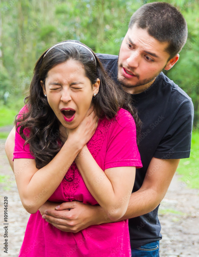Young woman choking with man standing behind performing heimlich maneuver, park environment and casual clothes