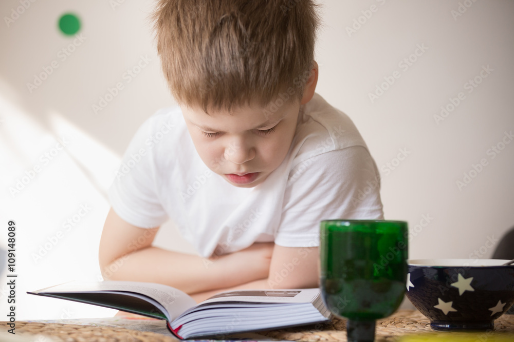 Cute kid boy reading a book while having his meal. Breakfast with reading.Child sitting at the table, studying and drinking and eating. Learning. Cute kid boy reading a book while having his meal. Breakfast with reading.Child sitting at the table, studying and drinking and eating. Learning.