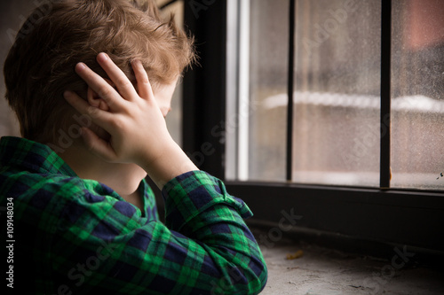 Little boy standing behind the window in sad mood. Sad Teenager looking in the Window and closing his ears with hands. Unhappy child in a plaid shirt. Alone at home. Upset.