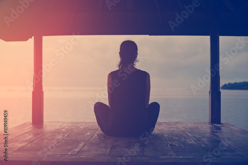 Silhouette of woman sitting and meditating near ocean at sunrise (intentional sun glare)