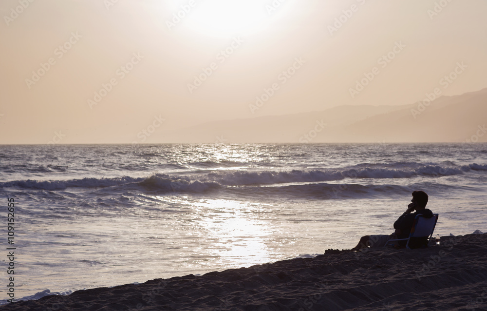 The silhouette of a man sitting on a sun lounger by the ocean. Light and shadow, a man rests during sunset on the beach.