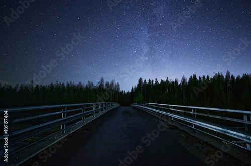 A road leading into night sky full of stars and visible milky way. A Bridge and dark forest on the foreground.