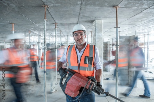 Fotografie Mid adult male builder holding building equipment  on busy construction site