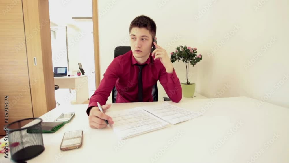 Young office worker sitting at his desk, talking using smartphone, taking notes; real people, real life; steadicam shot Young office worker sitting at his desk, talking using smartphone, taking notes; real people, real life; steadicam shot
