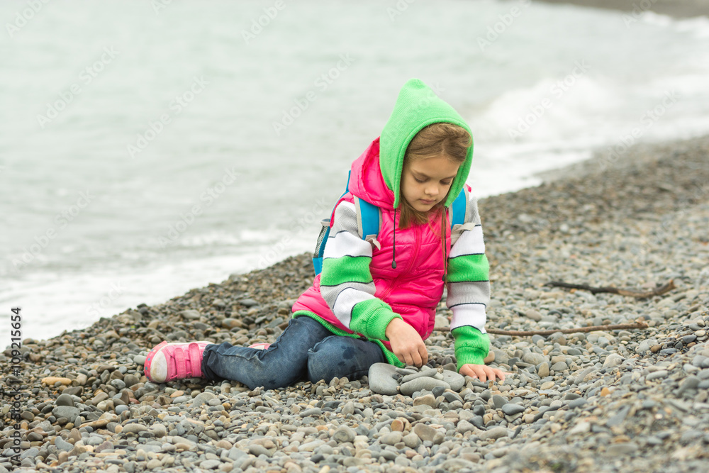 Seven-year girl sitting on a pebble beach in the warm clothing and with enthusiasm plays with stones Seven-year girl sitting on a pebble beach in the warm clothing and with enthusiasm plays with stones