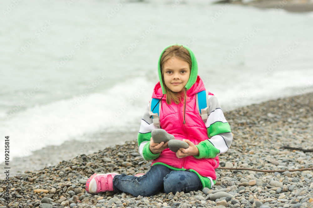 Seven-year girl sitting on a pebble beach in the warm clothes and looks in the frame Seven-year girl sitting on a pebble beach in the warm clothes and looks in the frame