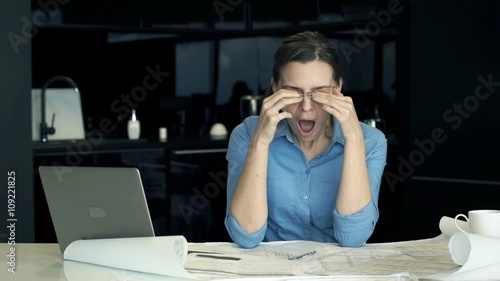 Sleepy, tired architect yawning over blueprints by table in the kitchen
