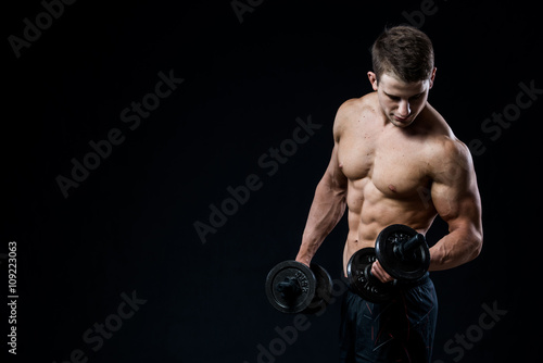 Handsome power athletic man training pumping up muscles with dumbbells in a gym. Fitness muscular body isolated on black background. Looking to his biceps.