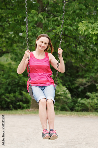 Portrait of happy smiling young middle age woman girl in red tshirt and jeans shorts on swing on backyard playground outside on summer day, lifestyle © anoushkatoronto