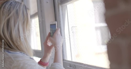 Over the shoulder view of beautiful young blonde woman using a smart phone to take pictures through a window in an urban hallway with brick walls. Hand held, slow motion, recorded in 4K at 60fps.
