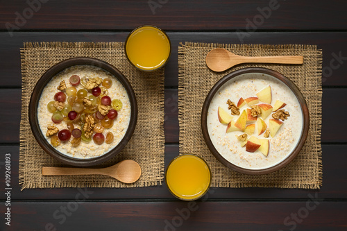 Oatmeal porridge with grapes, apples and walnuts in rustic bowls, glass of juice and wooden spoon on the side, photographed overhead on dark wood with natural light