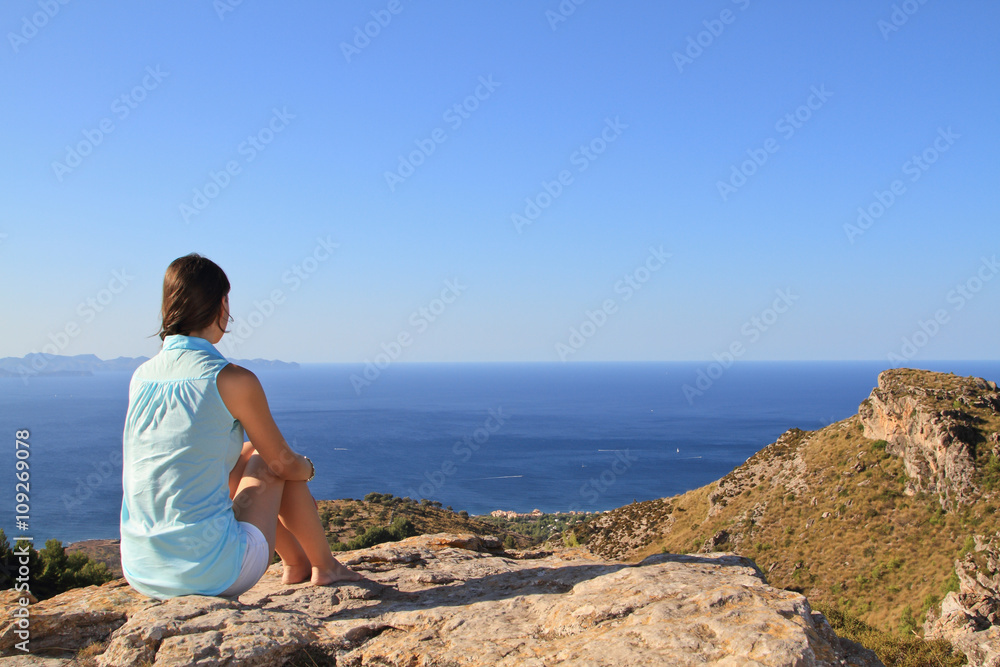 Contemplative beautiful girl sitting on the rocks in front of the mediterranean sea meditating. Blue and orange tones on the picture. This photo transmit calm, meditation, relax, holidays.