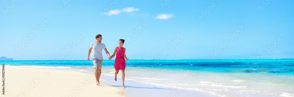 Panorama summer vacation couple walking on beach. Young adults having fun together enjoying their holidays travel in perfect getaway in sunny tropical destination with pristine turquoise ocean water.
