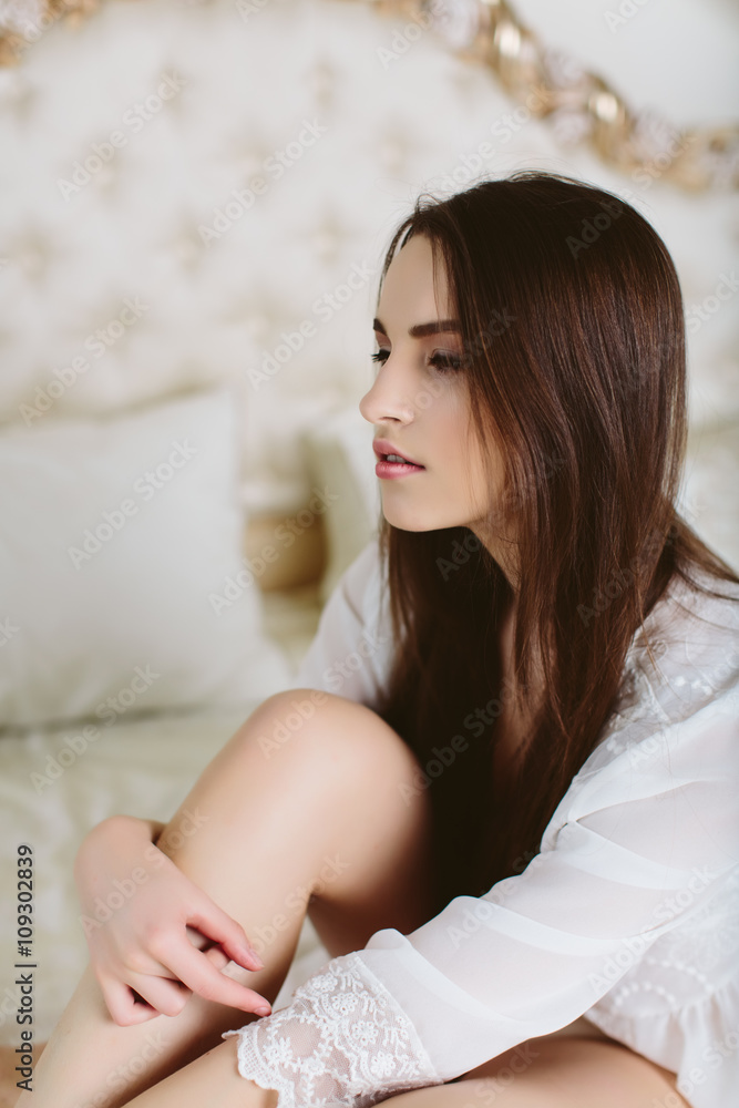 Model brunette woman in white translucent cloak sitting on bed and posing in boudoir room