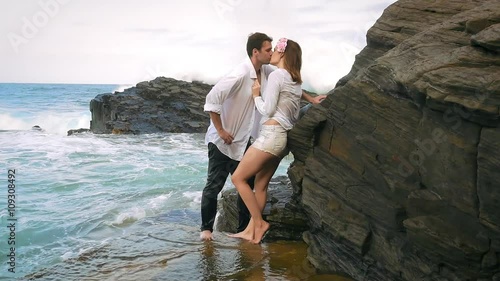 Young couple leaning against rocks kissing with waves crashing behind them
