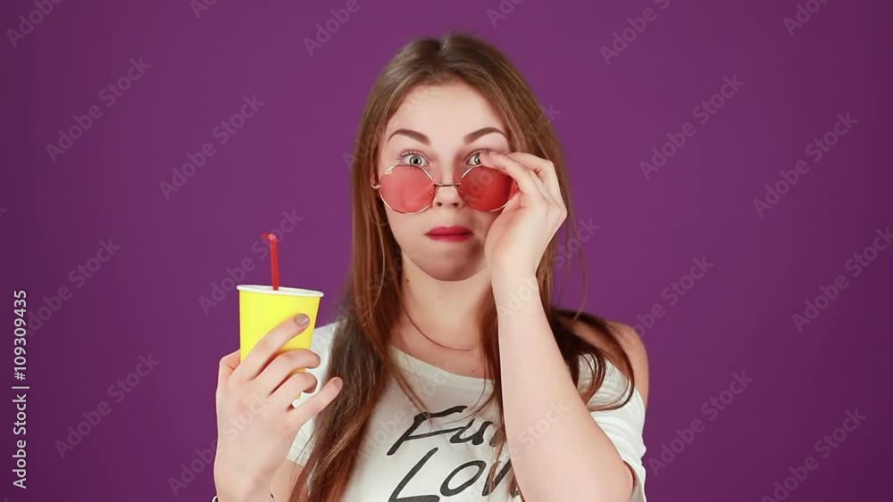 Stylish hipster girl with yellow paper cup puts on pink round glasses winks and smile Stylish hipster girl with yellow paper cup puts on pink round glasses winks and smile
