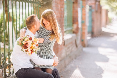 Close up fashion romantic portrait of young pretty couple kissing and hugs on the street at evening bright sunlight. Blonde gril with flowers boquet © nataliakabliuk