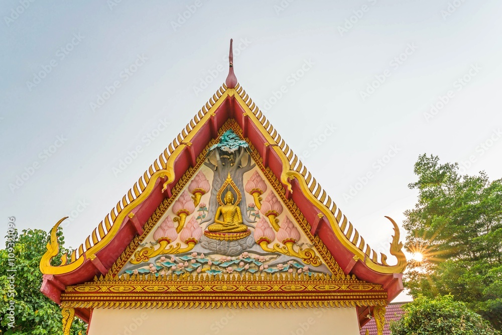 Sculpture of Buddha on public temple gable, triangle roof with sunray and trees, blue sky background Sculpture of Buddha on public temple gable, triangle roof with sunray and trees, blue sky background