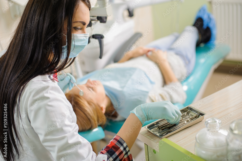 Young beautiful female dentist doing first check-up for female patient at the dental office. Doctor holding dental tools wearing mask and gloves. Medical equipment. Young beautiful female dentist doing first check-up for female patient at the dental office. Doctor holding dental tools wearing mask and gloves. Medical equipment.