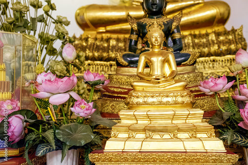 Close Up Of Small Golden Buddha Statue In Lotus Position With Flowers At Wat Pho, Bangkok, Thailand. Buddhism. Praying, Meditation. Religious Buddhist Symbol