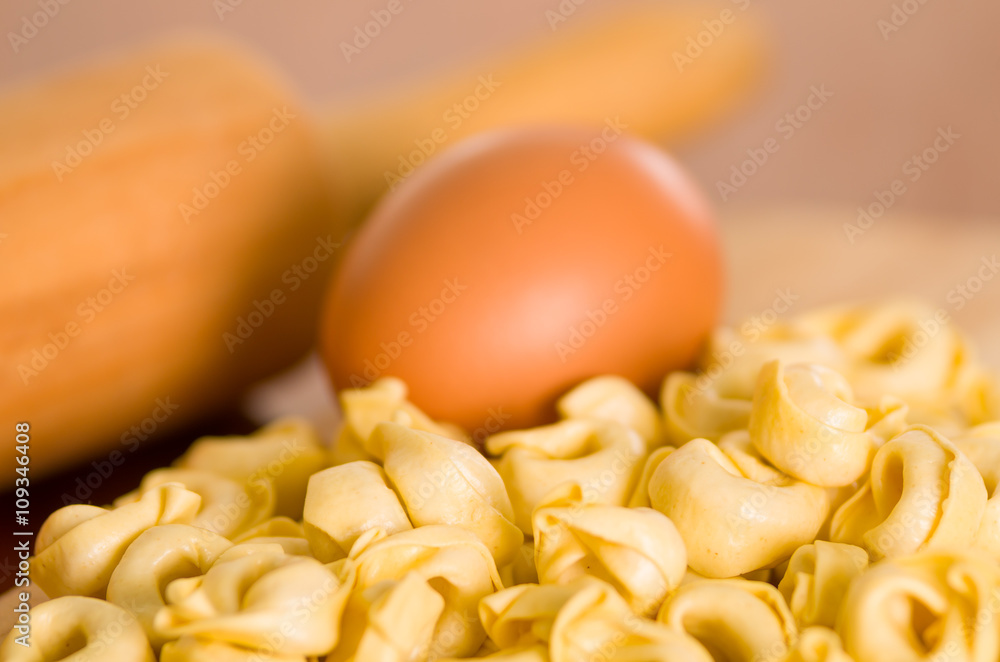 Closeup tortellinis piled up next to an egg and blurry rolling pin background