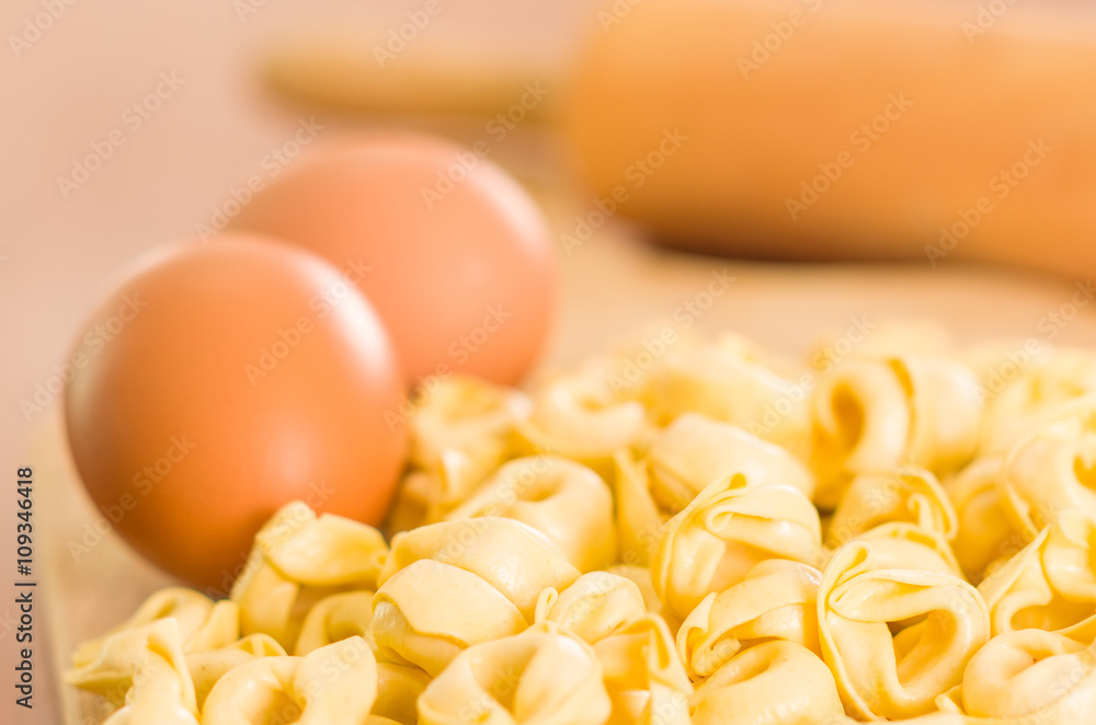Closeup tortellinis piled up next to an egg and blurry rolling pin background