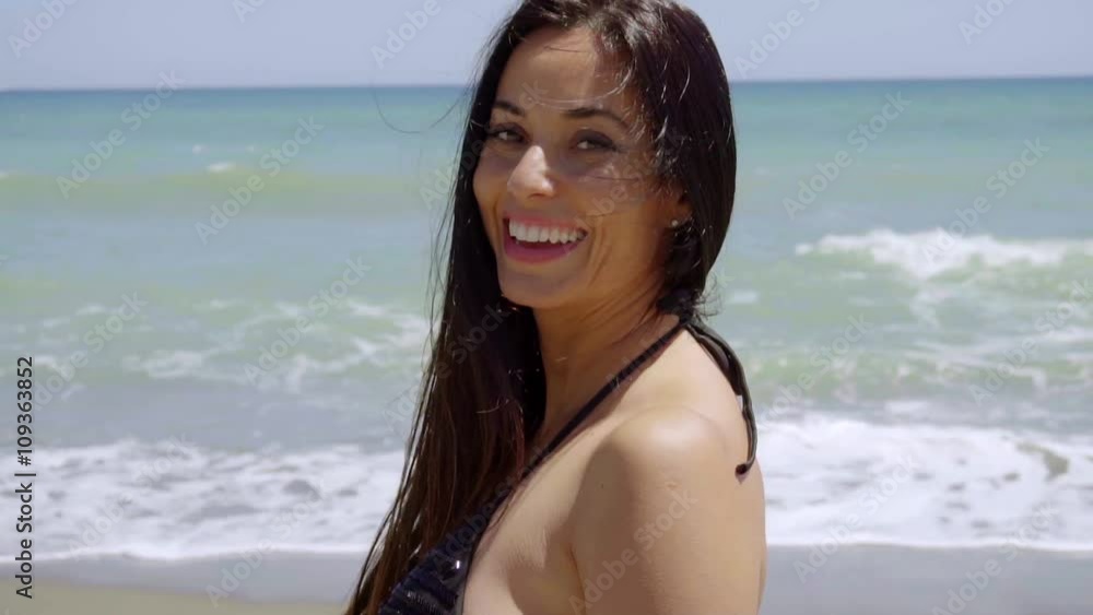 Gorgeous happy woman in a swimsuit standing on a tropical beach turning to look at the camera with a lovely warm friendly smile Gorgeous happy woman in a swimsuit standing on a tropical beach turning to look at the camera with a lovely warm friendly smile