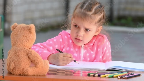 Five-year girl with enthusiasm draws a teddy bear sitting in front of her and a voluntary sticks out his tongue photo