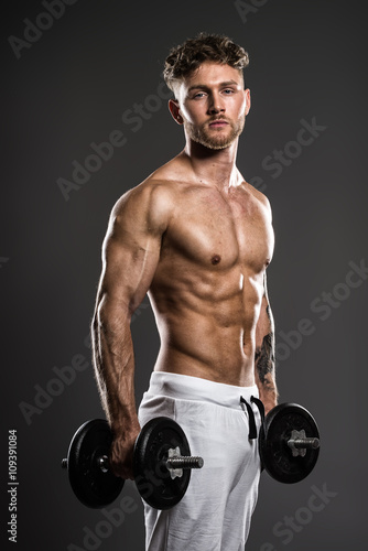 Handsome young bodybuilder training withdumbbells, on grey