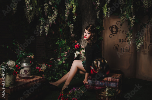 Magic portrait of romantic beautiful girl with wavy hair, red lips, art dress, holding rose flower, sitting on books. Female in scenery of Alice in Wonderland. Fashion fairy tale about princess