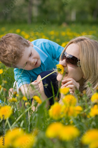 Happy family playing on green grass in spring park. A young mother is lying on the grass in a meadow of dandelion flowers with her son, smiling and smelling a flower.