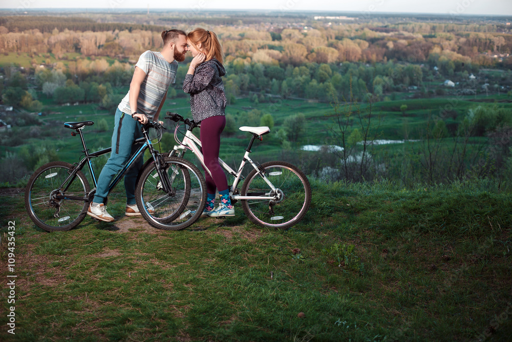Obraz premium Beautiful young couple in love walking with bicycles, kissing and hugging. Stand near the bikes. The concept of active rest