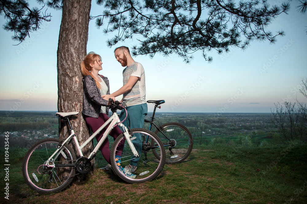 A young couple in love standing under a tree and hugging at sunset, standing next to the bike
