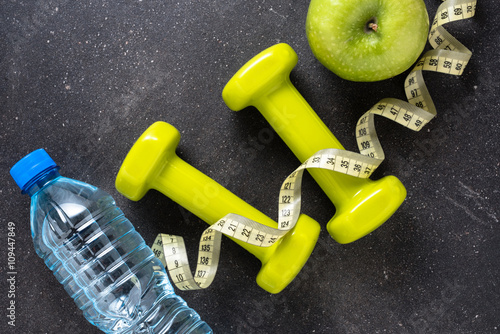 Fitness concept with dumbbells, water, tape measure and green apple on dark background
