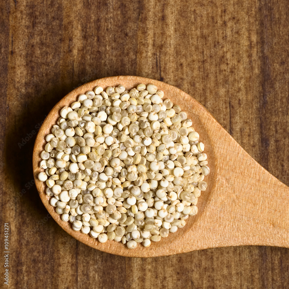 Raw organic white quinoa (lat. Chenopodium quinoa) grains on small wooden spoon, photographed overhead on dark wood with natural light.. Raw organic white quinoa (lat. Chenopodium quinoa) grains on small wooden spoon, photographed overhead on dark wood with natural light..